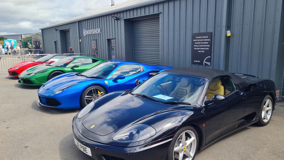 Four supercars lined up outside grey building. One car is black, one is blue, one is green and one is red.