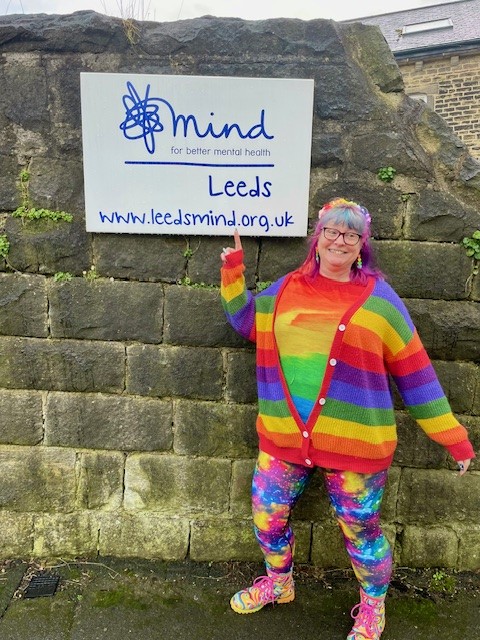 Mary is dressed in a bright rainbow striped cardigan and t-shirt, with rainbow tie-dyed leggins. She is posing with the Leeds Mind sign at Clarence House, Horsforth