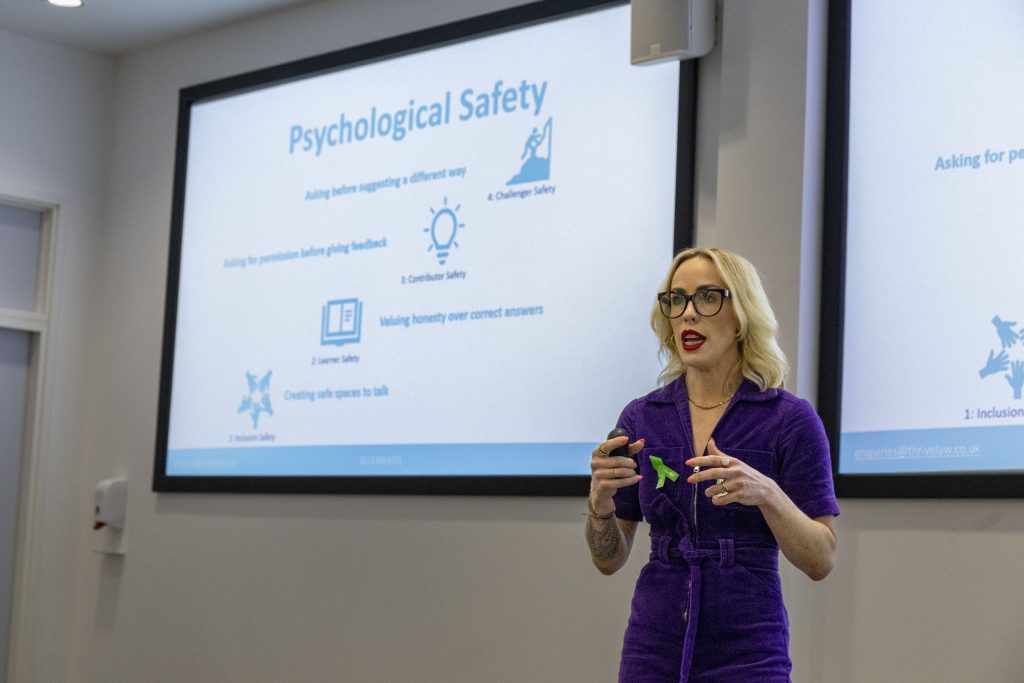 A woman wearing glasses stood in front of projector slides giving a presentation.