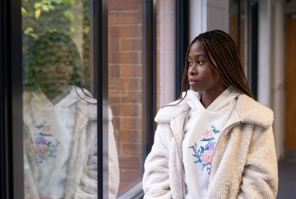 A young woman looks pensively out the window, and we see her reflection in the glass